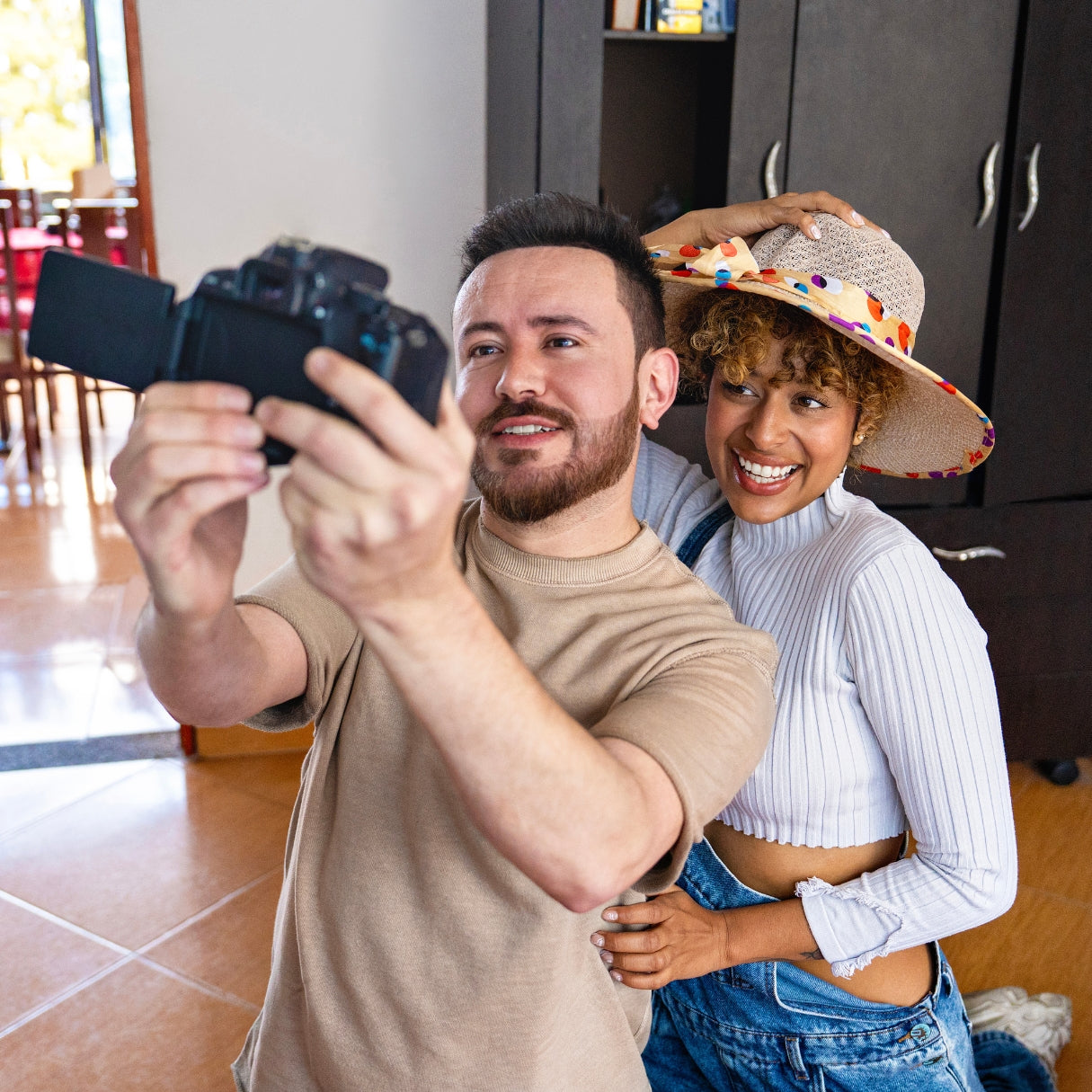 Man taking a selfie with a woman in a colorful hat in a kitchen.